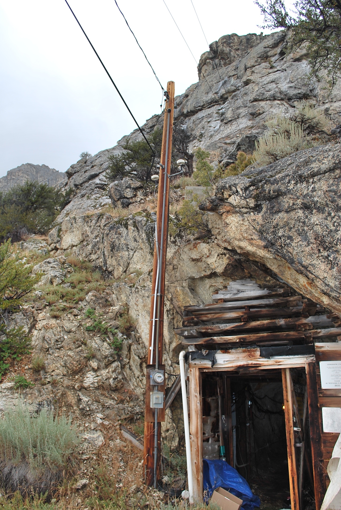 A telephone looking pole with a wooden building built into the rocky mountain behind it. 