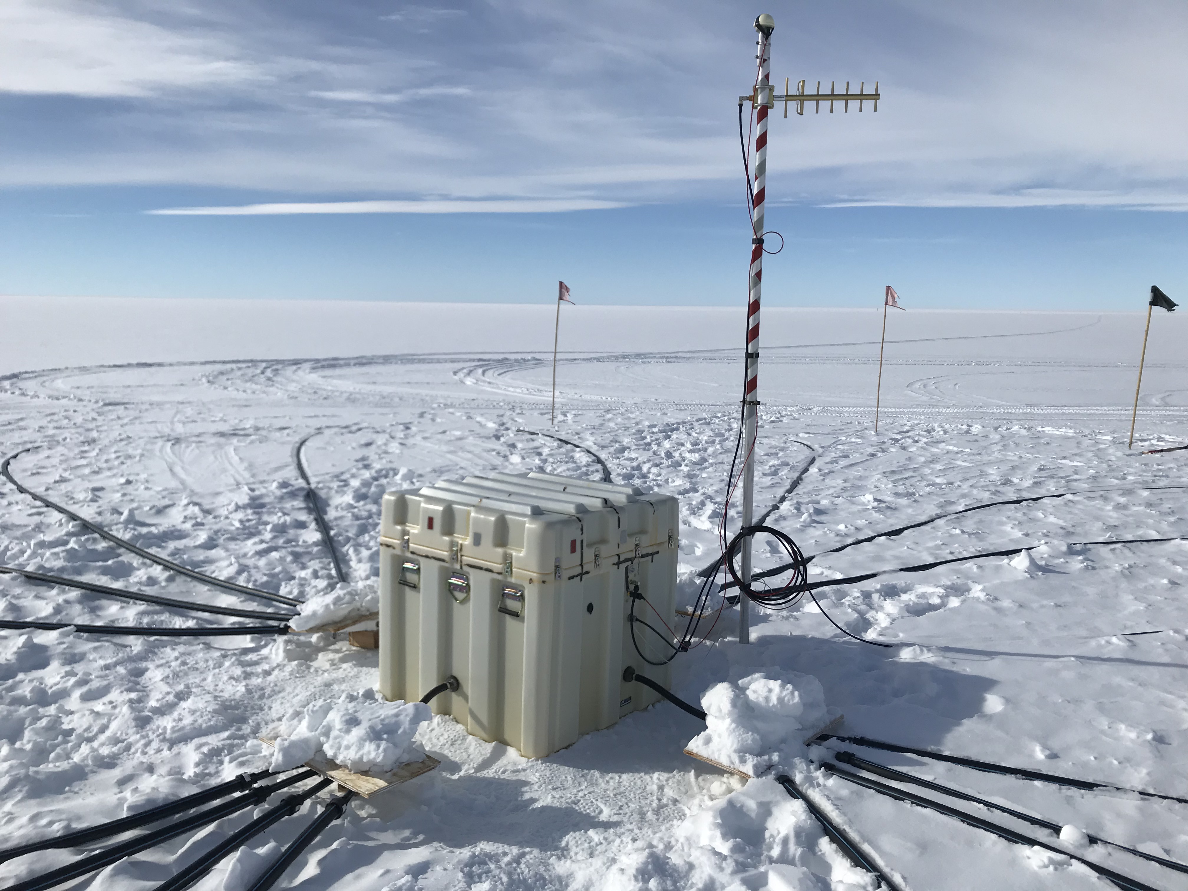 A large box, next to striped pole, with thick black wires coming out in all directions. Flags can be seen in the distance. Snow everywhere. 