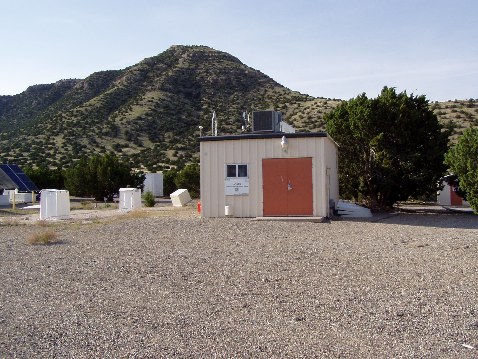 A small tan building with white boxes to its left outside in the sun. A green mountain is displayed behind it.